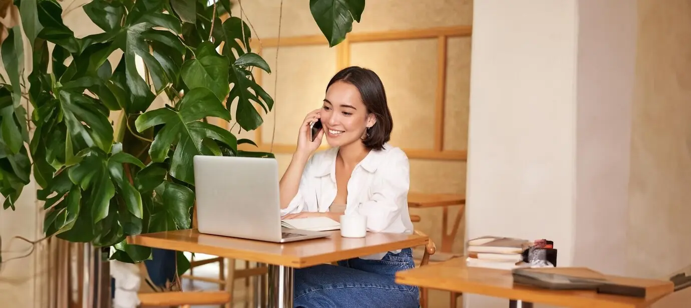 Joven gerente y propietaria de una cafetería, sentada con un portátil, contestando llamadas telefónicas y sonriendo amablemente.