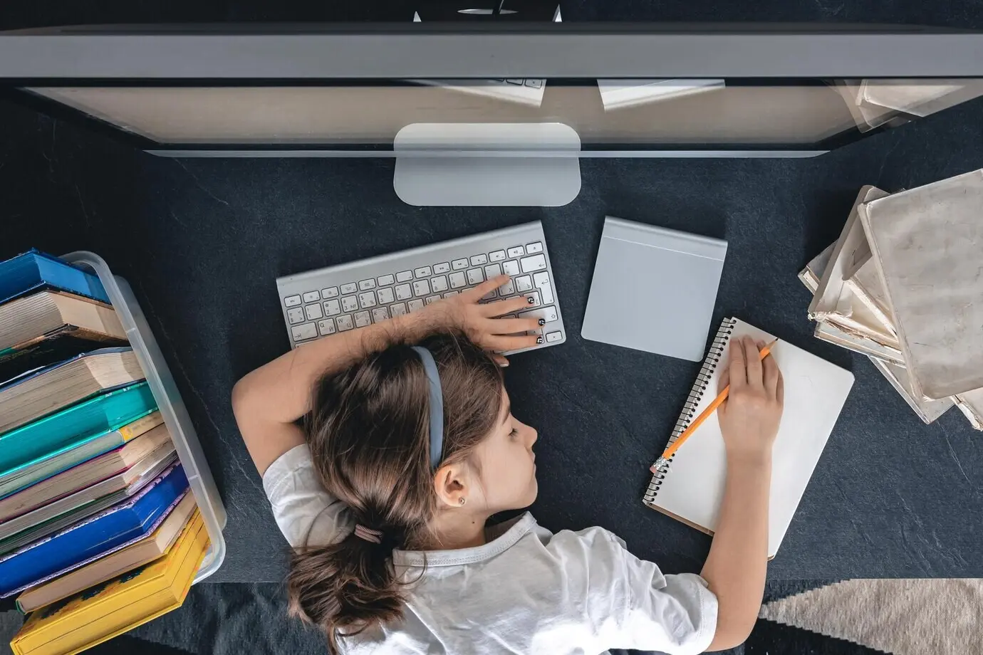 Una chica cansada está acostada sobre una mesa frente a una computadora, con libretas y libros.